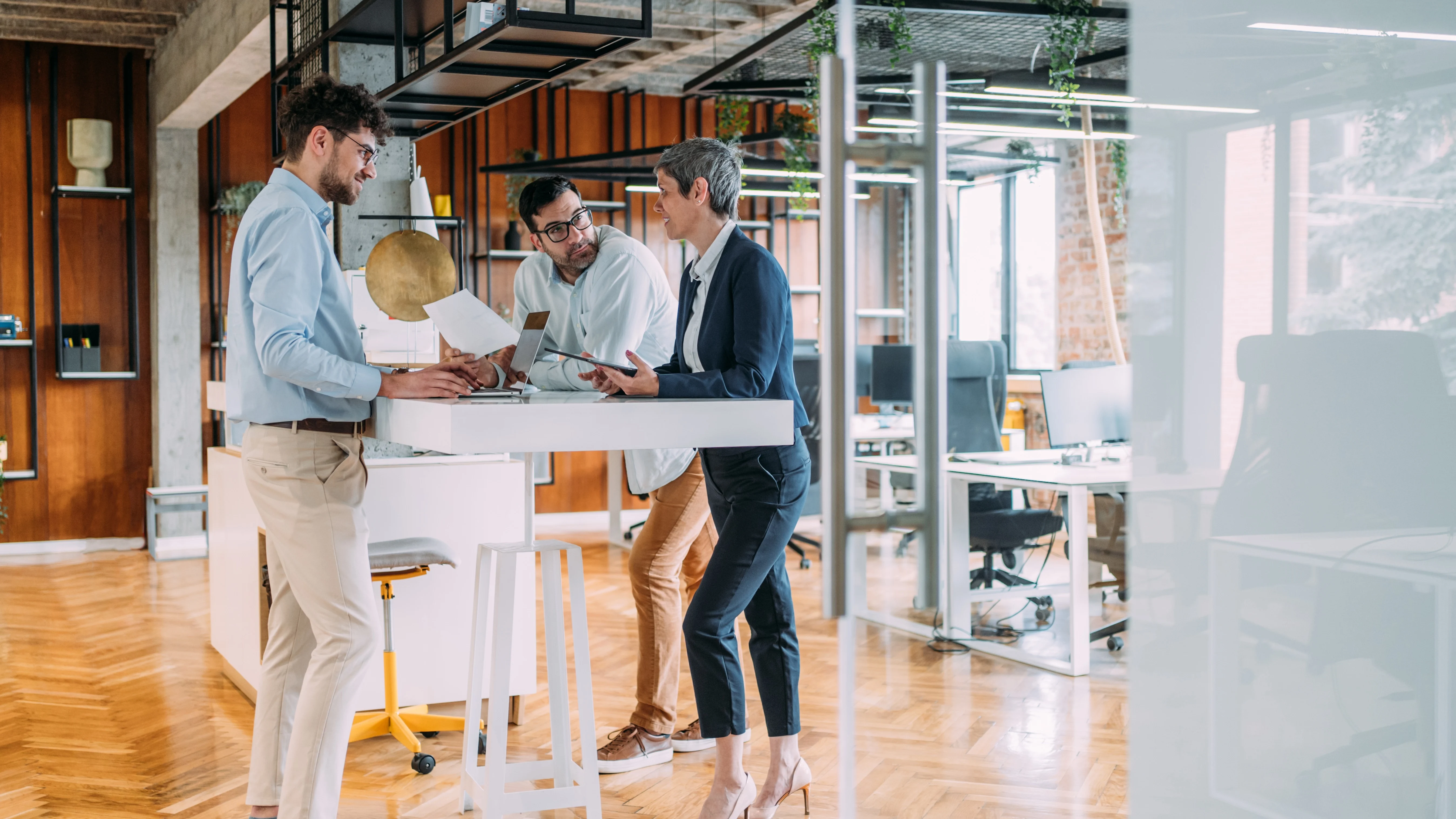 Three professionals collaborating at a standing desk in a modern office with wooden floors and industrial design elements.