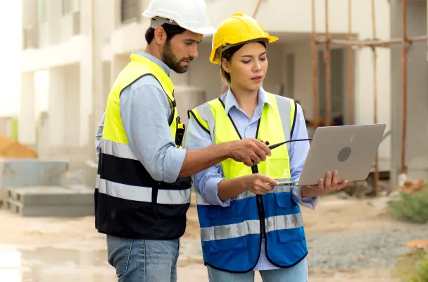 Two construction workers in high-vis vests analyse a laptop screen on a construction site. 