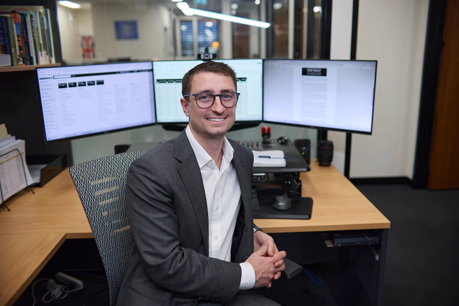 A smiling man in a grey blazer and glasses sits at a desk with three computer monitors in a modern office.
