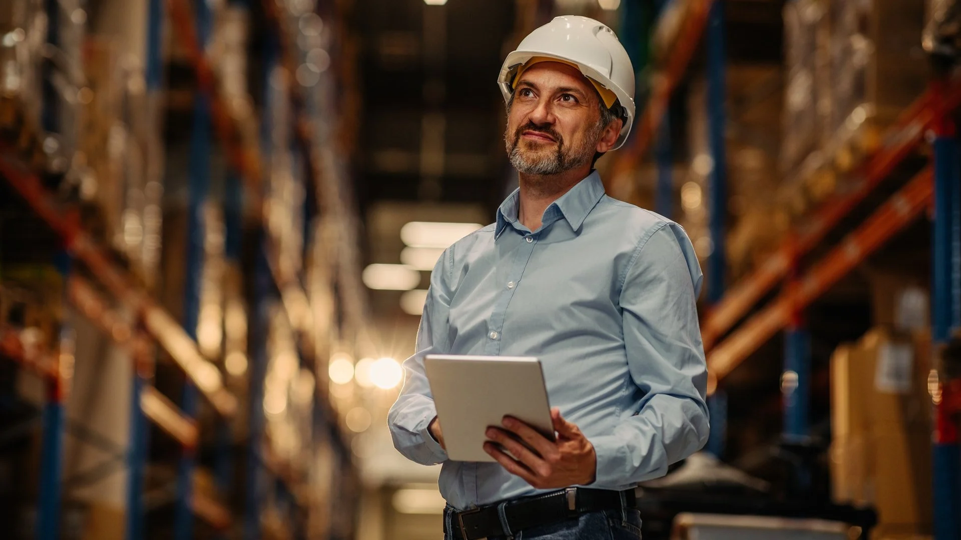 Worker in white hard hat holding tablet while looking up in warehouse with tall storage racks and ambient lighting.