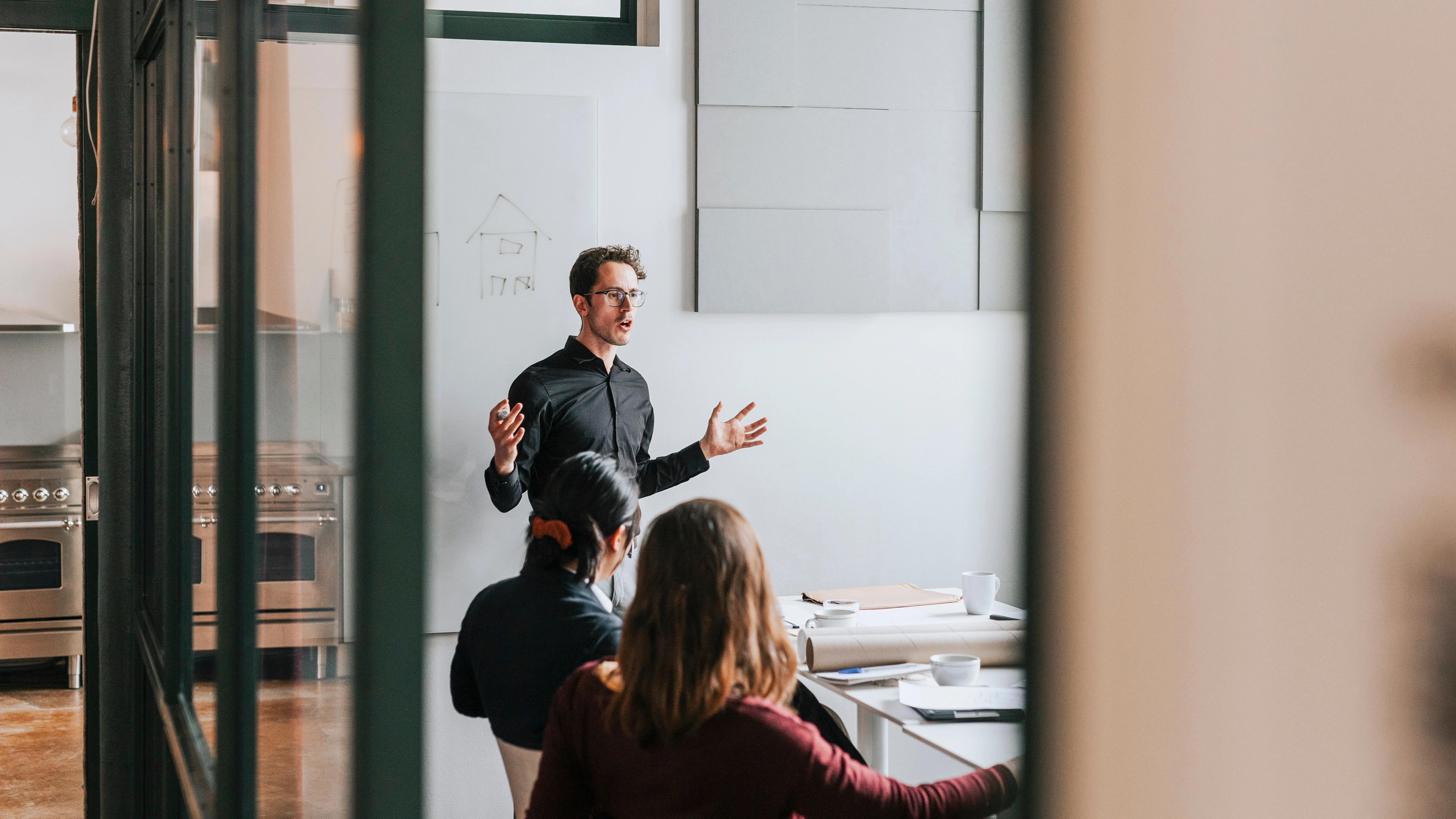 Person in black shirt presenting to small audience in modern office meeting room with whiteboard showing house sketch.