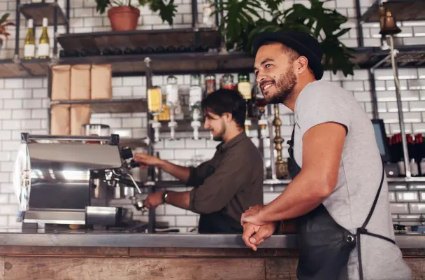 A waiter in a cafe leans on the countertop smiling while a barista makes coffee in the background.