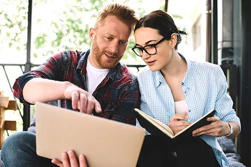 Two franchisees exchange notes while reading from a laptop. One takes notes in their notebook.