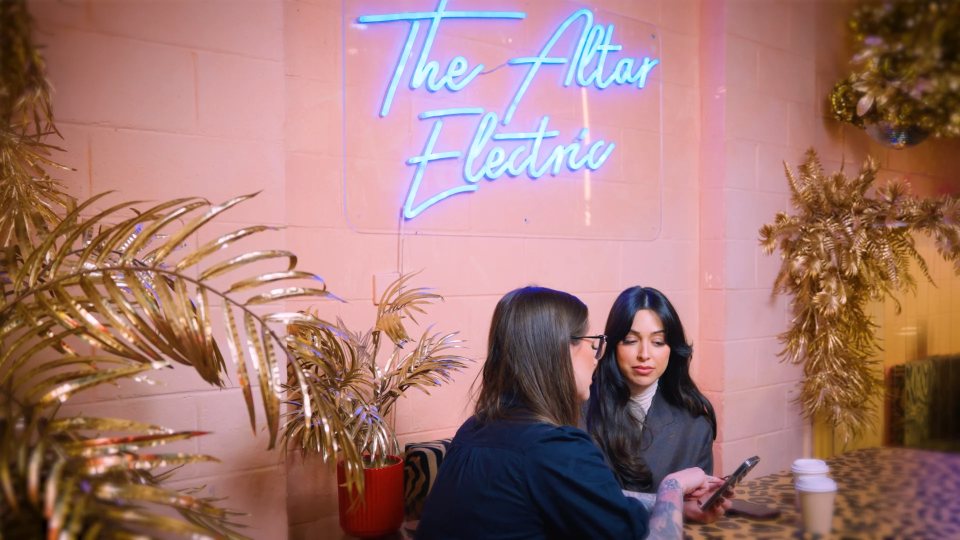Two people sitting at a table beneath blue neon sign reading "The Altar Electric" with decorative plants nearby.