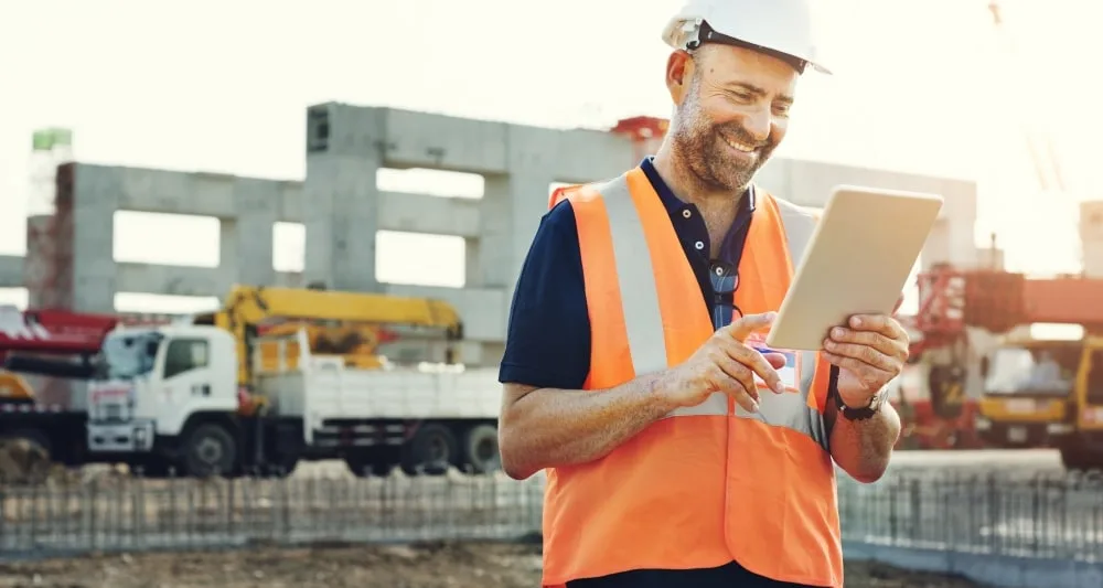 A construction worker at a building site, wearing a hard hat and high visibility jacket, interacting with a tablet computer whilst smiling.