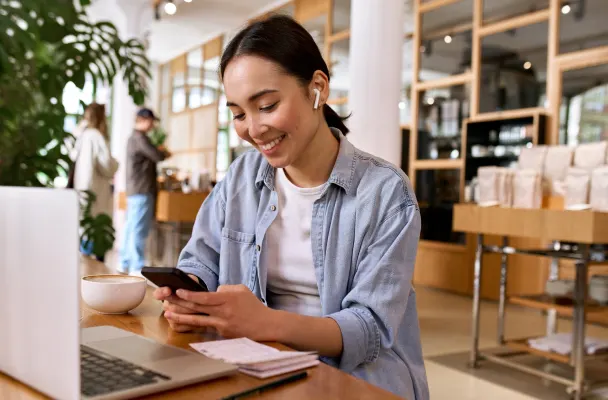 Student sits in a library with her laptop and notes in front of her while smiling at their phone.