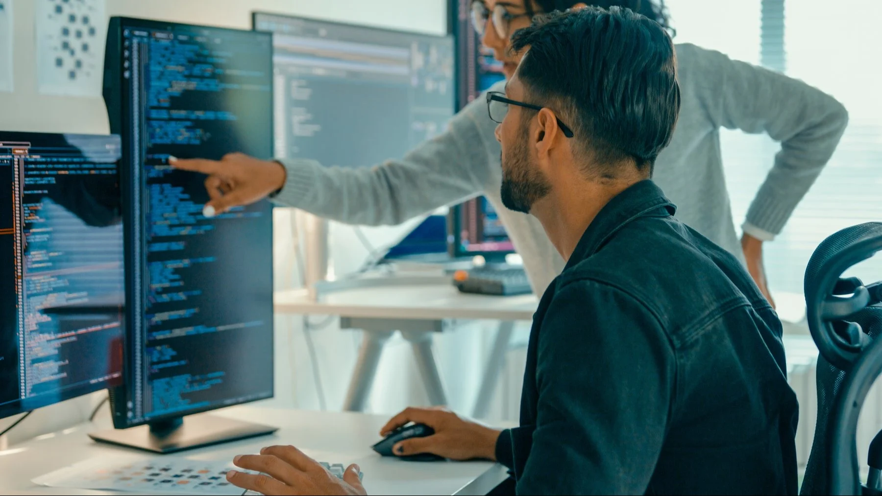 Two software developers collaborating on code, one seated at computer while colleague points at screen showing blue text.