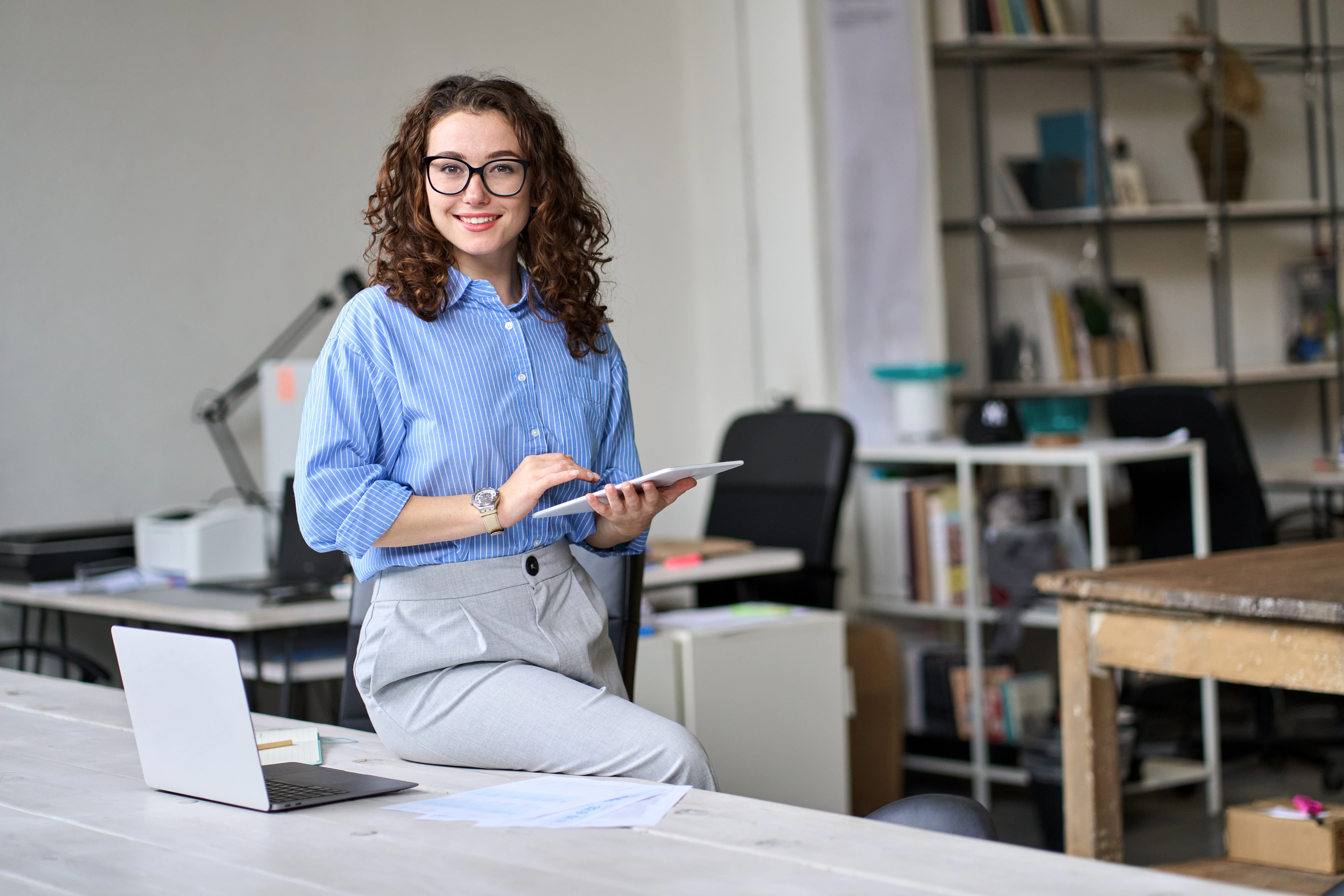 A young woman, sitting on a desk with a laptop in front of her and a tablet in her hand. She is looking directly at the camera and smiling wide. She is in a shared office space.