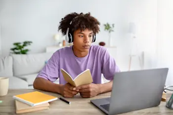 a teenage boy wearing headphones in a purple tshirt reading a book whilst looking at an open laptop.