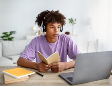 a teenage boy wearing headphones in a purple tshirt reading a book whilst looking at an open laptop.
