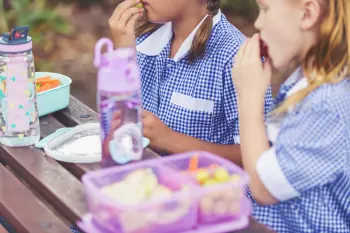 Two young girls in school uniform sit together on a bench eating packed lunches