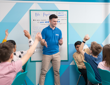 A tutor stands in front of a group of children with their hands raised to answer a question.