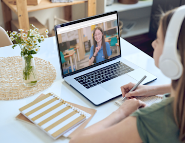 A teenage girl, wearing headphones, sits at a dining table with an open laptop in front of her. On the screen of the laptop an Explore tutor is smiling back at her.
