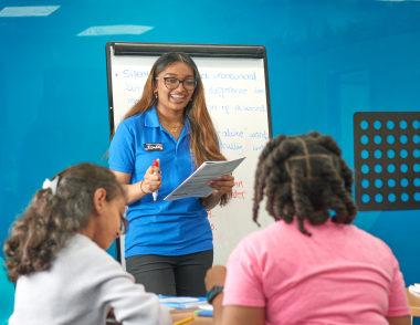 An Explore tutor stands in front of a whiteboard. There are two girls in the foreground working at a table.