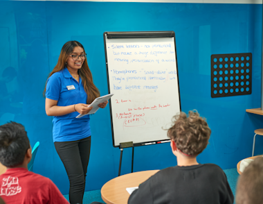 An Indian female tutor stands in front of whiteboard as she presents learning techniques to a class.