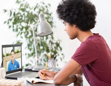 A young black teenager sits at a desk with an open laptop and notebook in front of him. On the screen there is an Explore tutor smiling back at him.