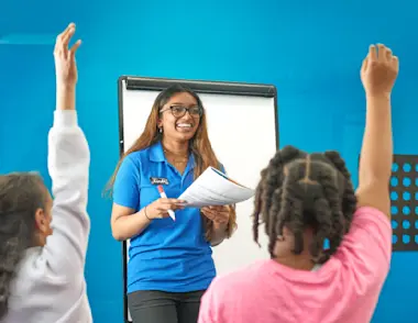 An Explore Learning tutor leading a class with children in foreground putting their hands up.