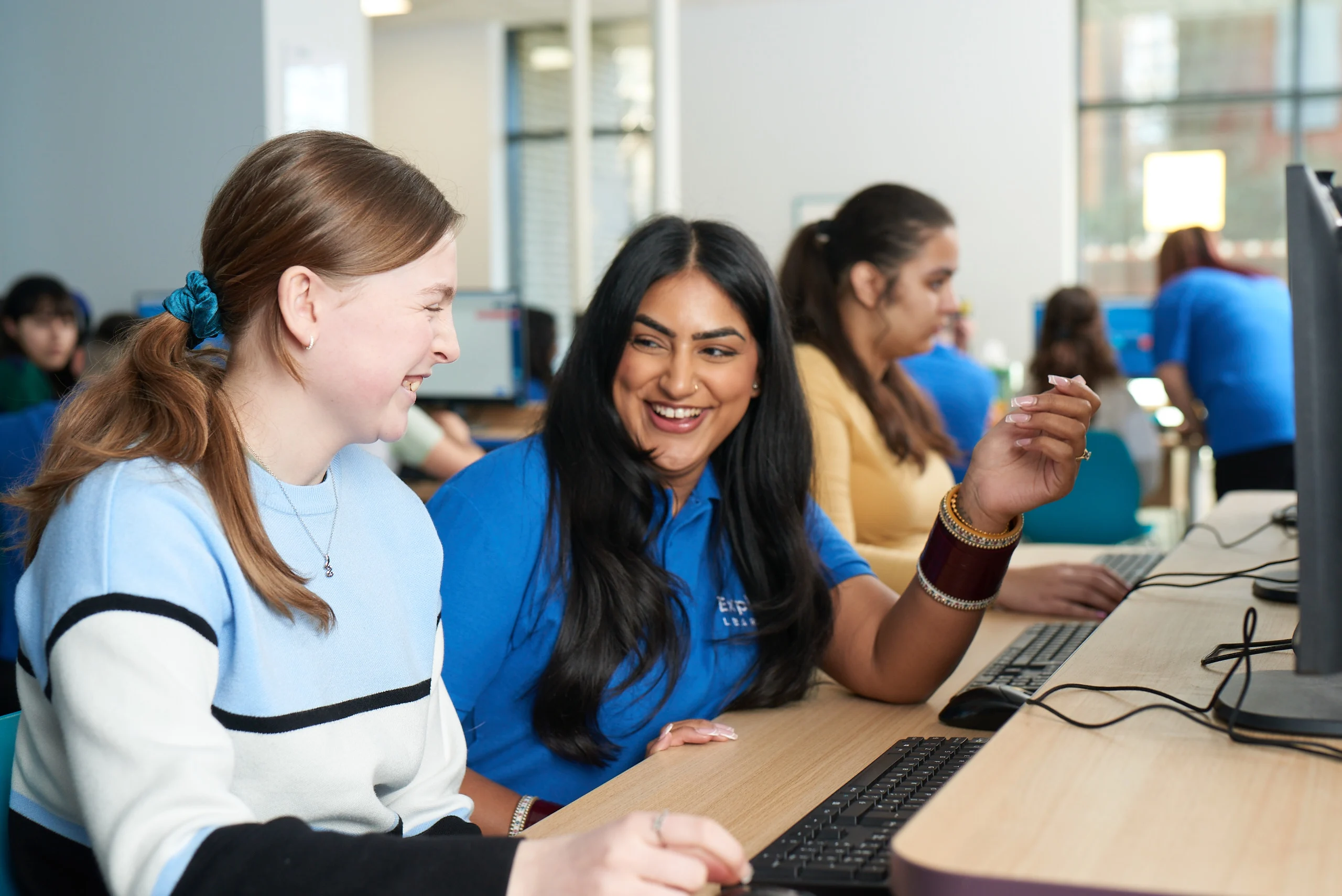 A female tutor laughs with a teenage girl as they bond while working together to cover GCSE topics on a computer.