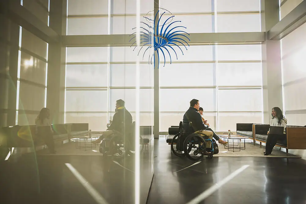 Several people sit together in a large, bright office. A man is sitting in a wheelchair, while a woman across from him sits on a bench with her laptop in her lap.