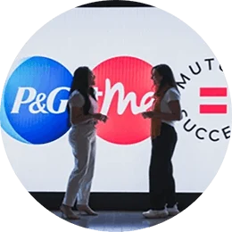 Two women face each other as they engage in conversation. They are standing at the end of a hallway, in front of a digital display of a blue, white and pink logo.