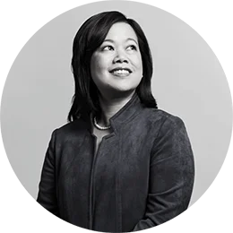 An Asian woman with shoulder-length black hair wears a dark suede jacket and a string of pearls. She smiles as she looks off in the distance. The image is black and white.