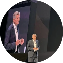 A white man in a grey business suit and white shirt stands on a stage. An illustrated of a brain, in the shape of a light bulb, and the white text that says "idea" are projected onto a large screen in the background.