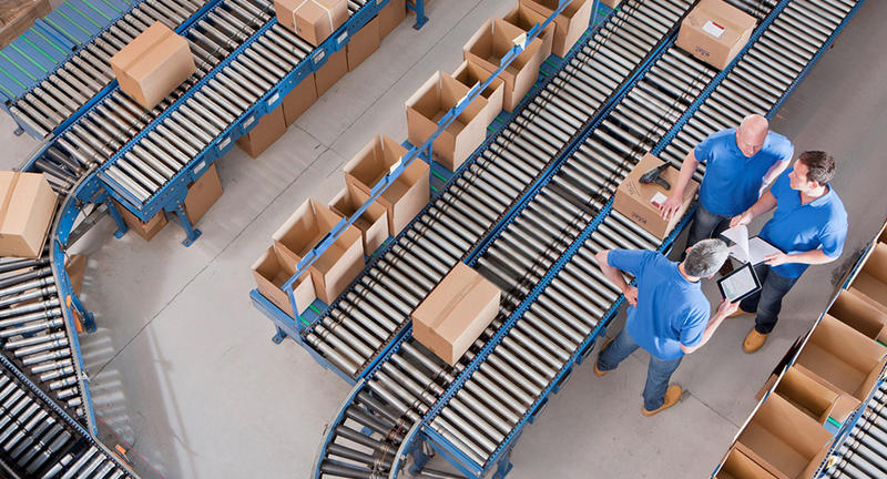 Men standing next to conveyor belts in a warehouse, looking at a tablet