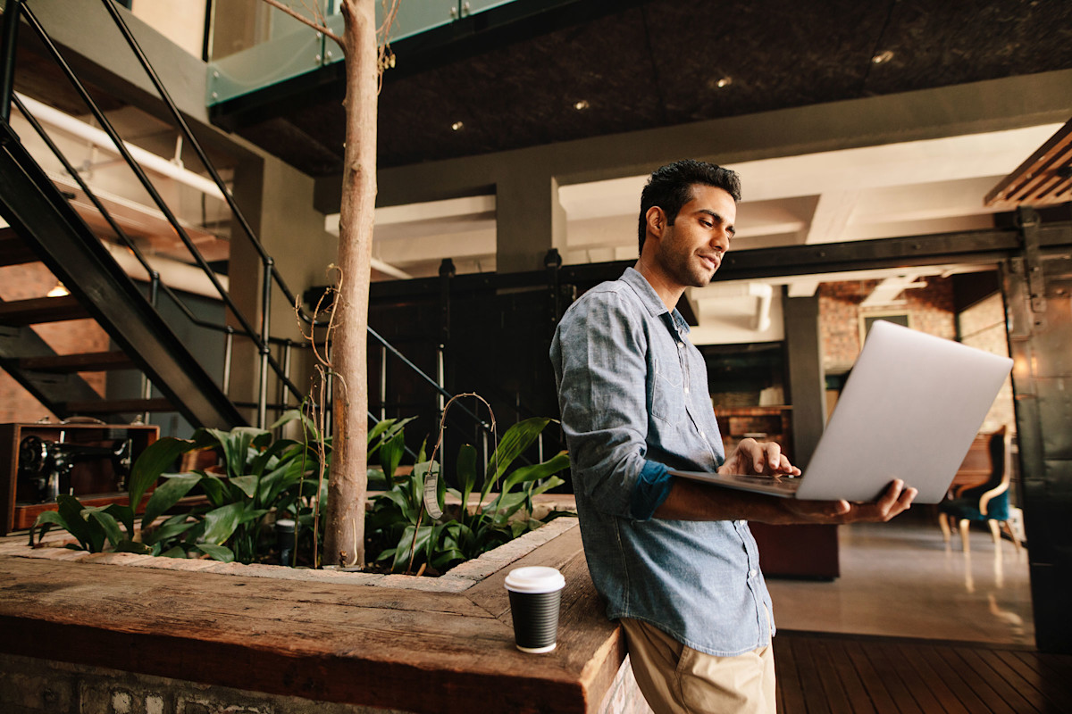 Man working on a laptop in a hallway with a cup of coffee. Plants and a stairway behind him.