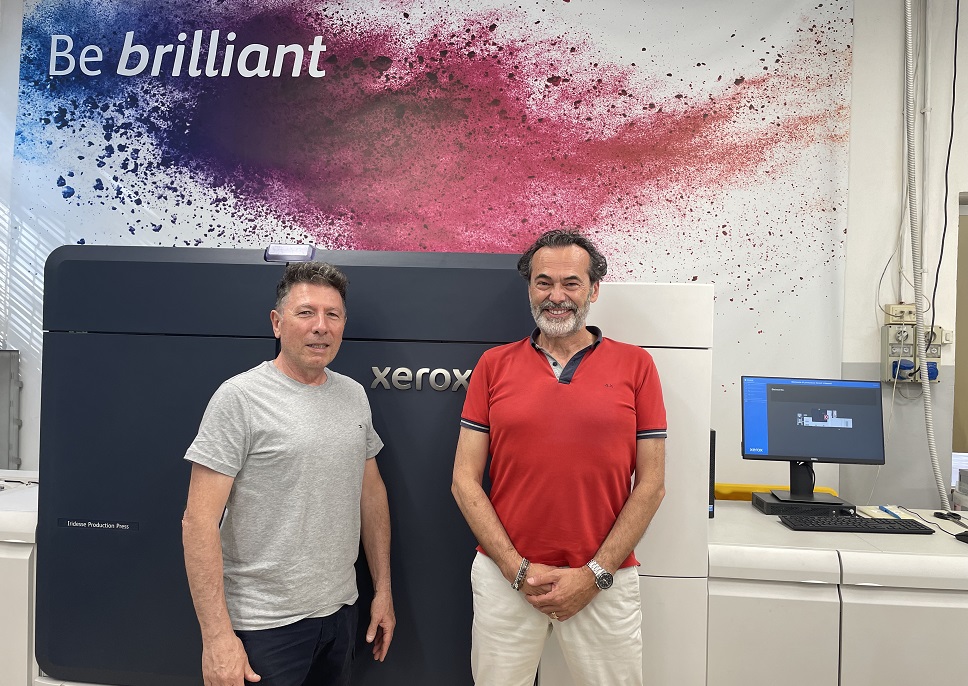Staff of Montagnani Print & Graphics in front of their Xerox Iridesse Production Press. A sign in the background says "Be brilliant" over blue and red ink droplets.