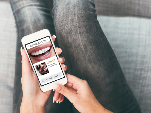 Picture of hands holding a smartphone with a webpage titled "Dental Jewelry" displayed along with a thumbnail of a smile with a dental gem adhered to a front tooth