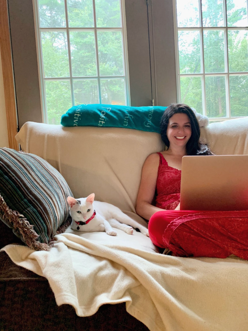 Luna sits beside Tamara on the black couch covered with a white blanket. Tamara smiles as she works on her laptop as Luna lay beside her. Luna's red collar matches Tamara's long red sundress.