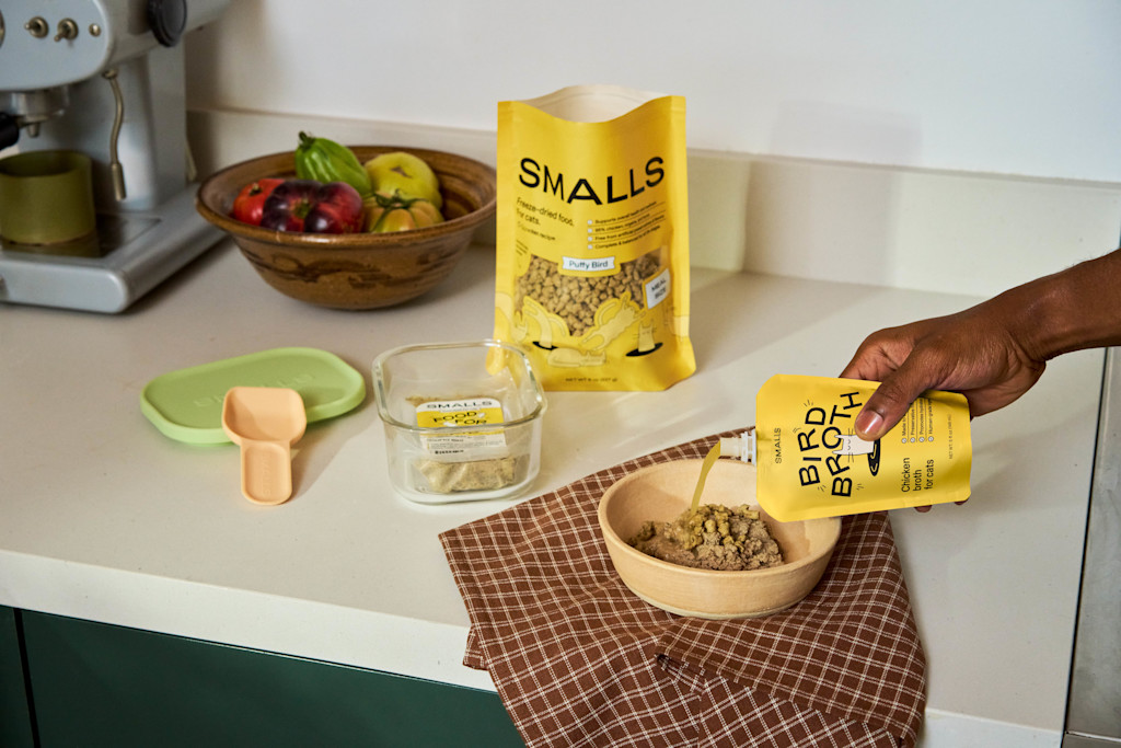 The image shows a kitchen countertop setup with a scene of preparing cat food using Smalls products. A hand is seen pouring liquid from a yellow packet labeled "BIRD BROTH" into a wooden bowl filled with cat food. On the counter, next to the bowl, there's a small brown checkered cloth. To the left of the bowl, a clear glass container with a green lid and a beige measuring scoop is visible, holding a small amount of food. A larger yellow bag labeled "SMALLS - Puffy Bird" is set upright in the background, suggesting it's another product for feeding cats. A bowl of colorful fresh fruits, including apples and tomatoes, sits towards the back of the counter. The kitchen has a light and clean aesthetic, with neutral colors, highlighting the natural ingredients of the Smalls products.