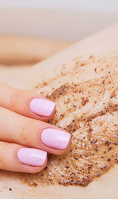 Mano con unghie rosa che applica uno scrub marrone sulla pelle, presentando un trattamento di bellezza per l'esfoliazione e la cura della pelle.
