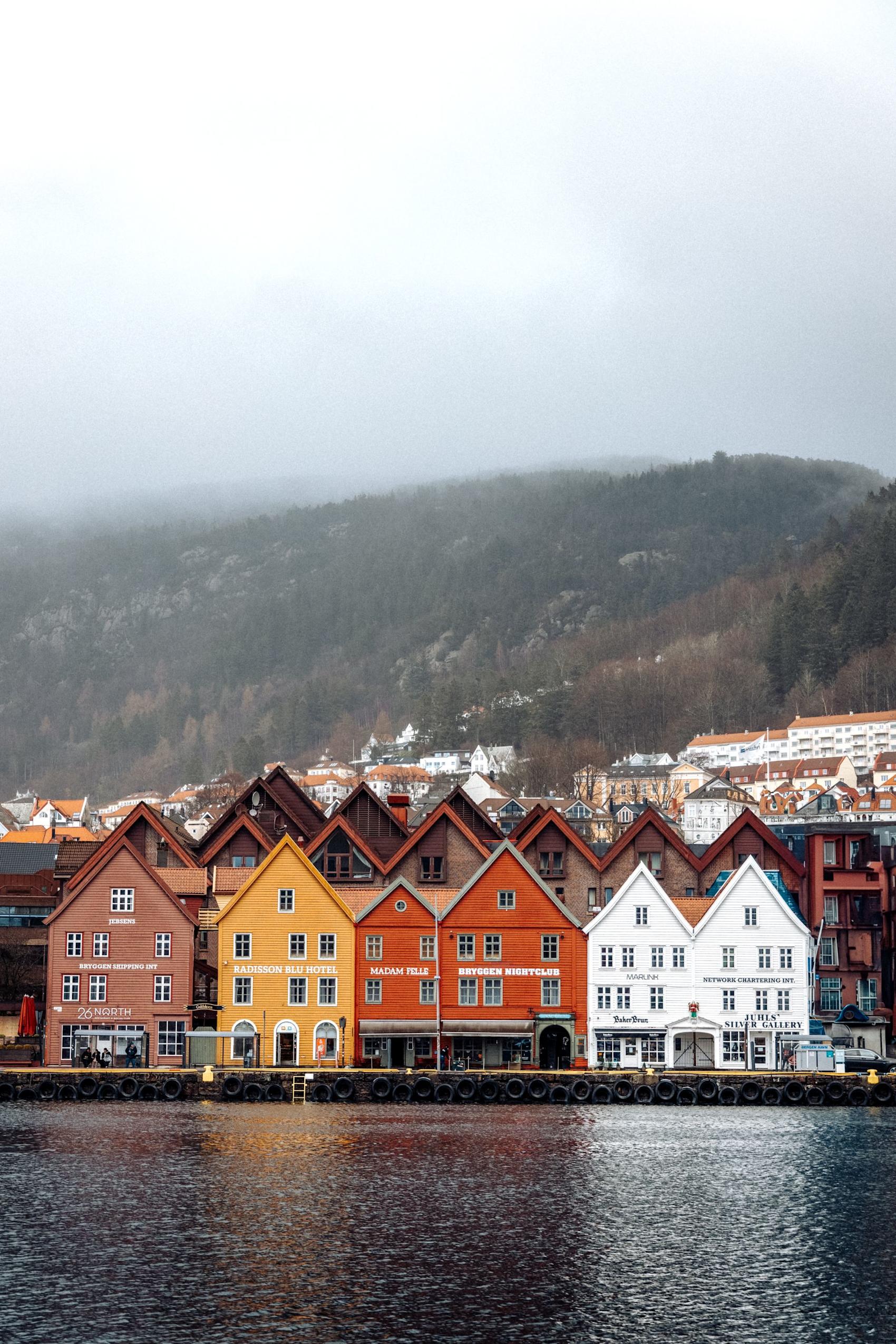 Fargerike trehus på Bryggen i Bergen ved vannet, med tåkete fjell i bakgrunnen på en overskyet dag. Foto: Vivek Tedla, Pexels.