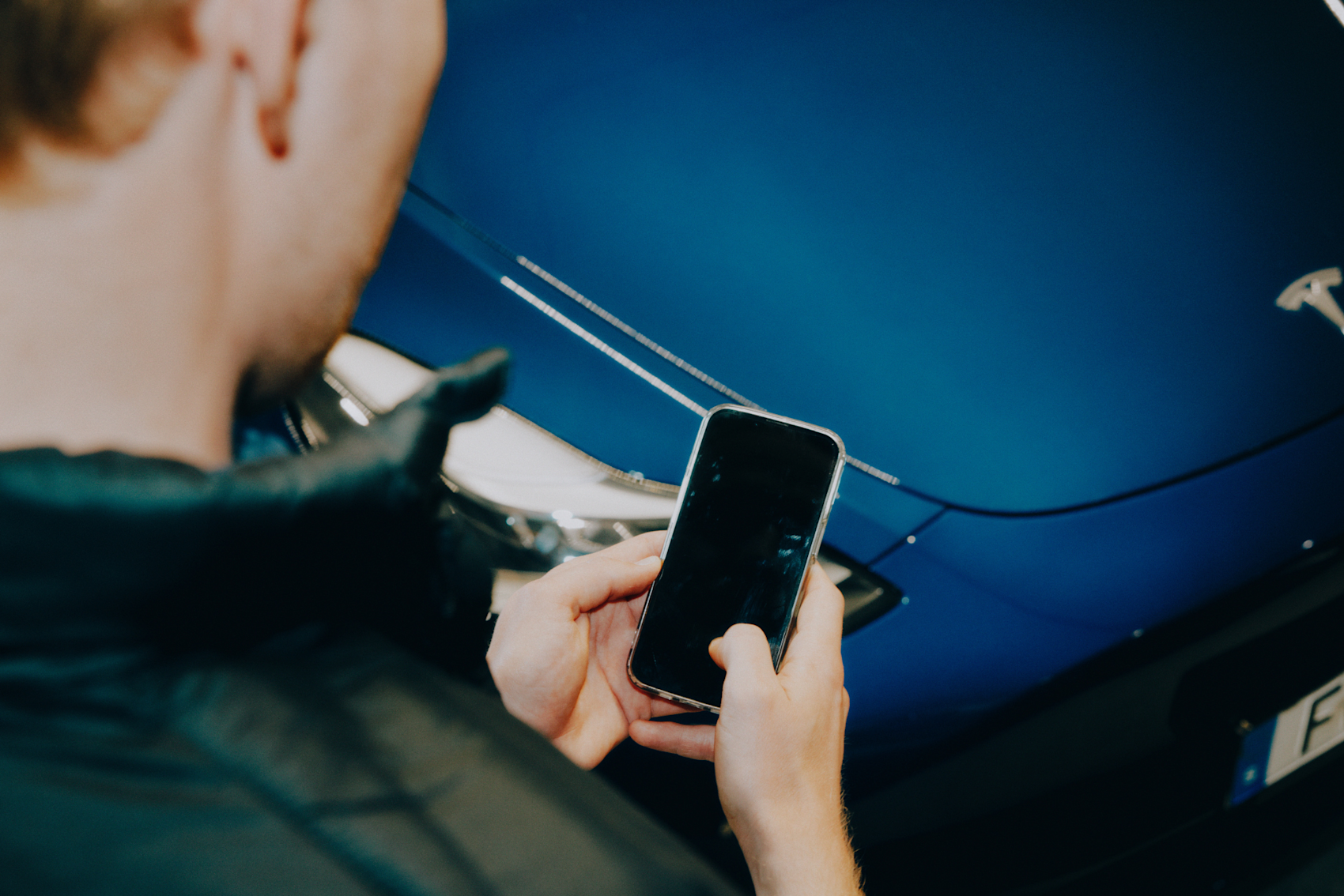 En mann holder en smarttelefon foran en blå bil, sett bakfra. Han ser ut til å fotografere eller inspisere bilen.