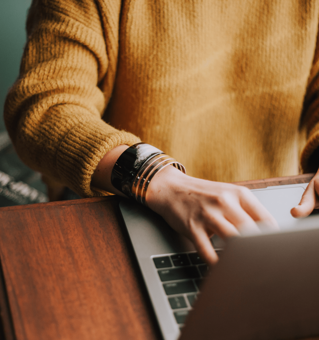 Woman in brown knit sitting at table typing on a laptop computer SaferVPN promo image of woman on laptop