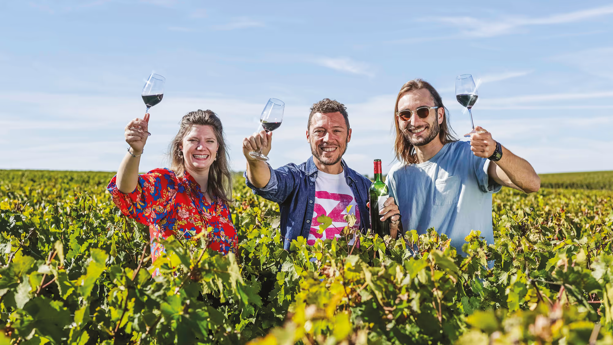 Three people standing among vineyard rows, raising glasses of red wine, with a half-drunk wine bottle held in the centre.