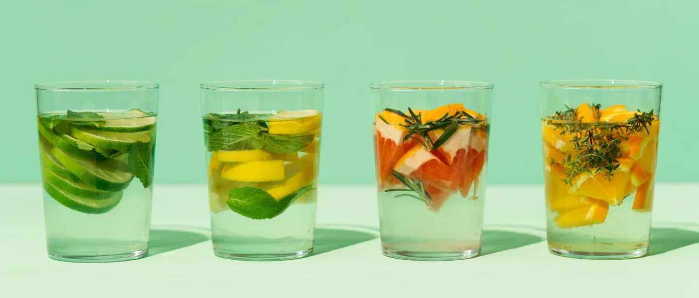 Four glasses of water with various fruits and herbs in them, such as sliced citrus, mint and rosemary).