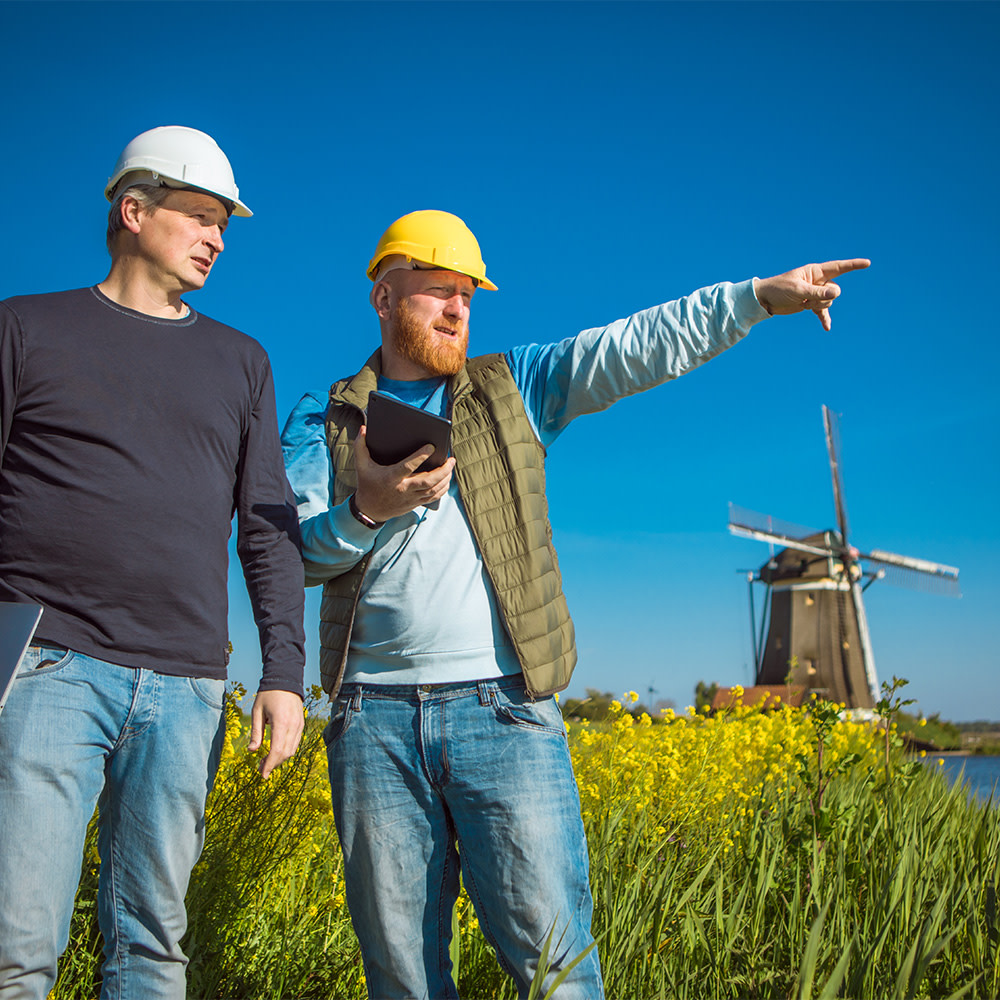 Zwei Männer mit Bauhelmen stehen vor einer Seenlandschaft mit einer Windmühle im Hintergrund.