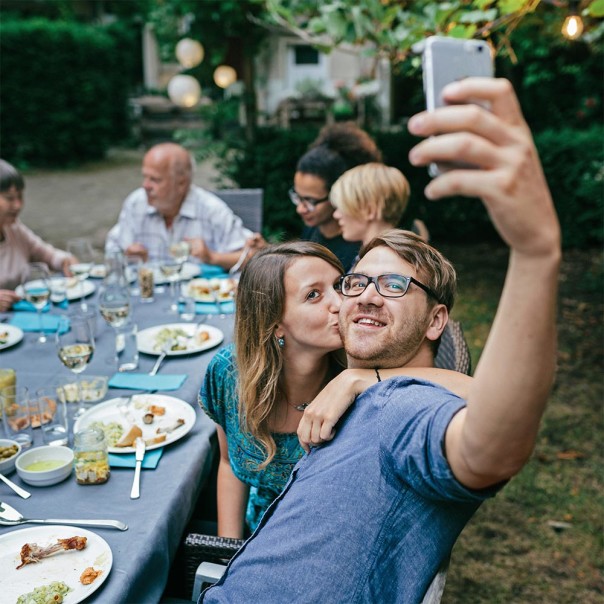 Mann sitzt an festlicher Tafel im Freien macht ein Selfie während Frau ihm auf die Wange küsst. Private Momente und Nachrichten bleiben auch mit und dank 5G privat. 