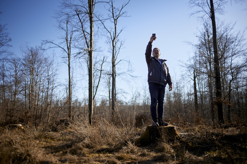 Mann steht im Wald auf einem Holzstumpf und misst mit einem Gerät in der Luft haltend Funk.