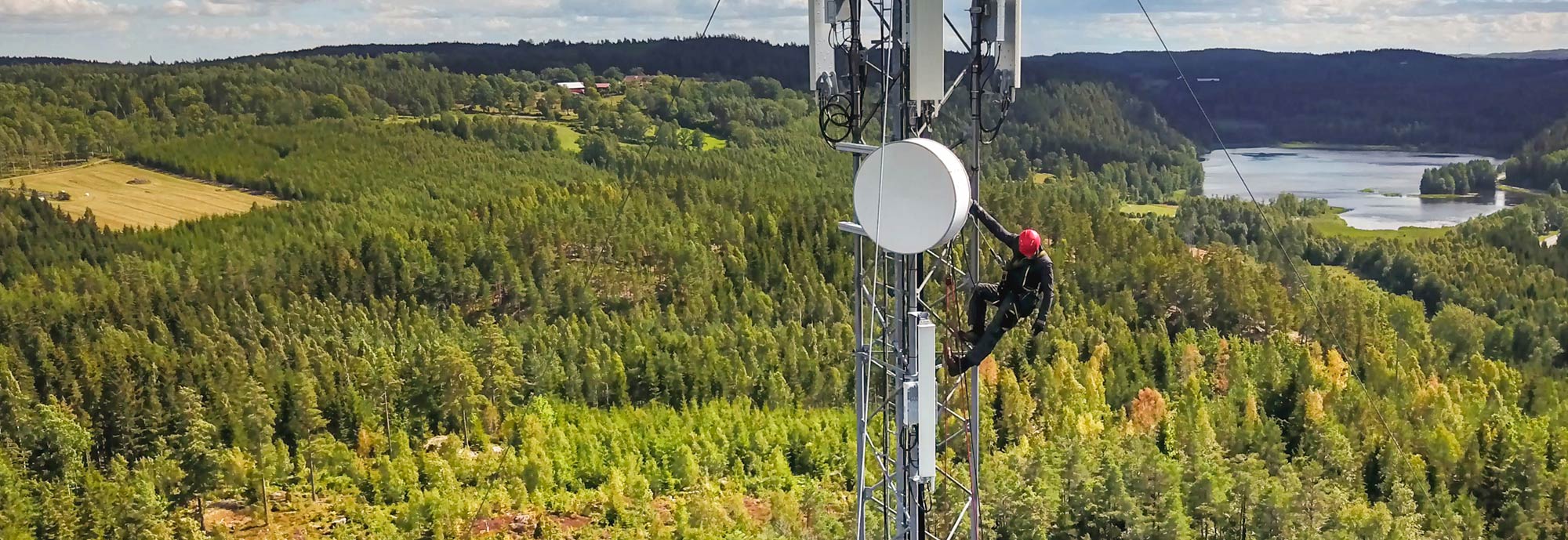 Ein Mobilfunk-Techniker klettert auf Sendemast vor einer Waldlandschaft. Ein 5G-Sendemast, der die umliegende Umgebung mit 5G Netz versorgt. 
