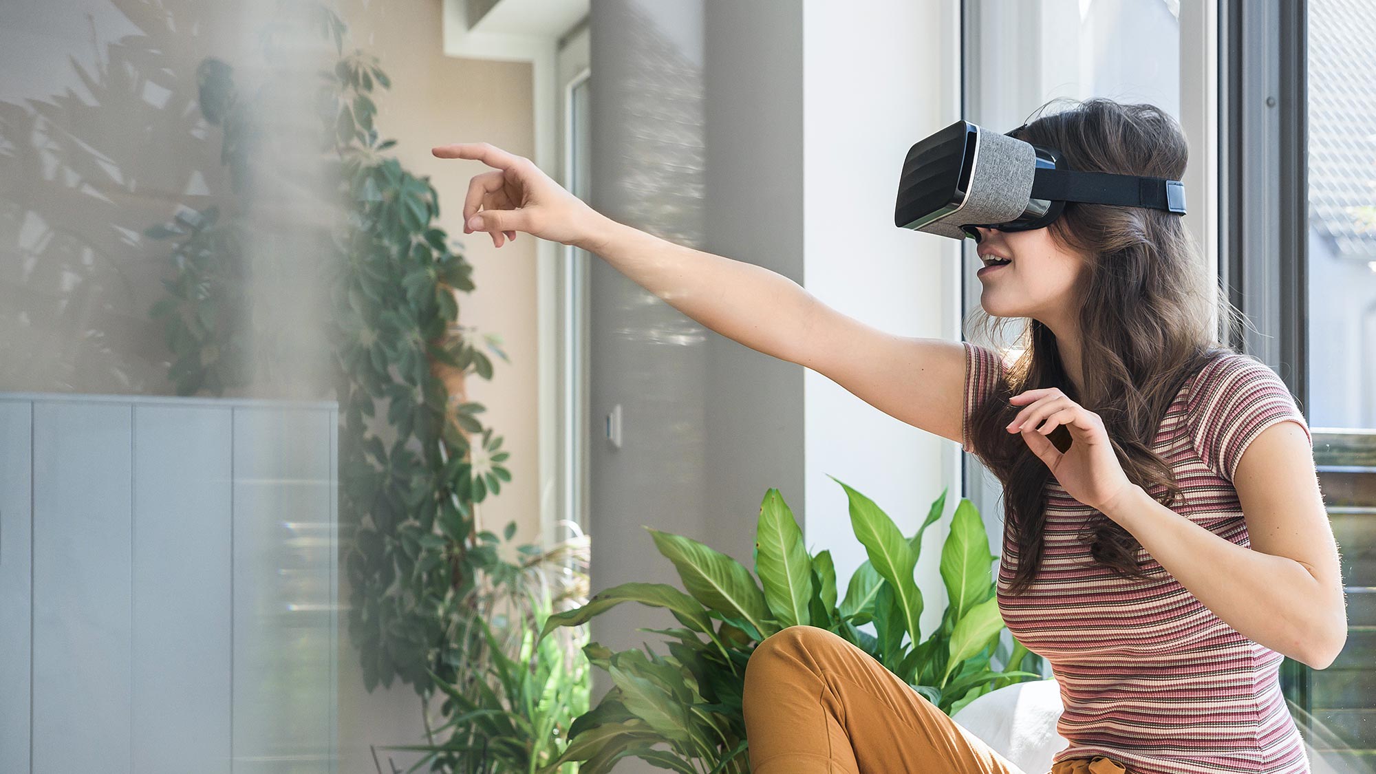 Frau sitzt auf Sofa und deutet mit dem Arm geradeaus während sie eine Virtual Reality Brille trägt. Mit dem 5G-Netz erkunden Nutzerinnen und Nutzer schnell und überall virtuelle Realitäten. 