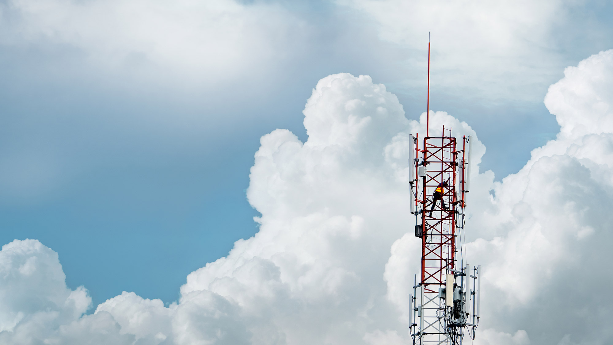 Mann steht auf einem Mobilfunkmast, der vor Wolken in den blauen Himmel ragt. 