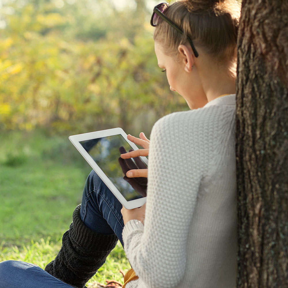 Frau sitzt mit Rücken an einem Baum gelehnt und bedient ein Tablet. Der neue 5G-Mobilfunk ist nicht nur für Kinder eine sichere Sache. 