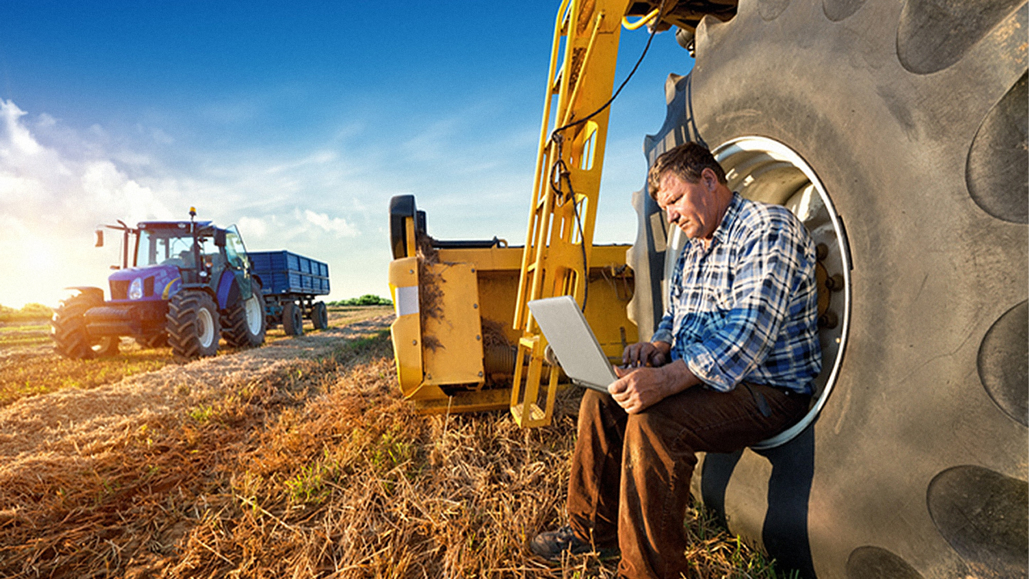 Ein Landwirt sitzt in einem großen Reifen und nutzt sein Laptop während der Arbeit auf dem Feld, im Hintergrund steht ein Traktor mit Anhänger.