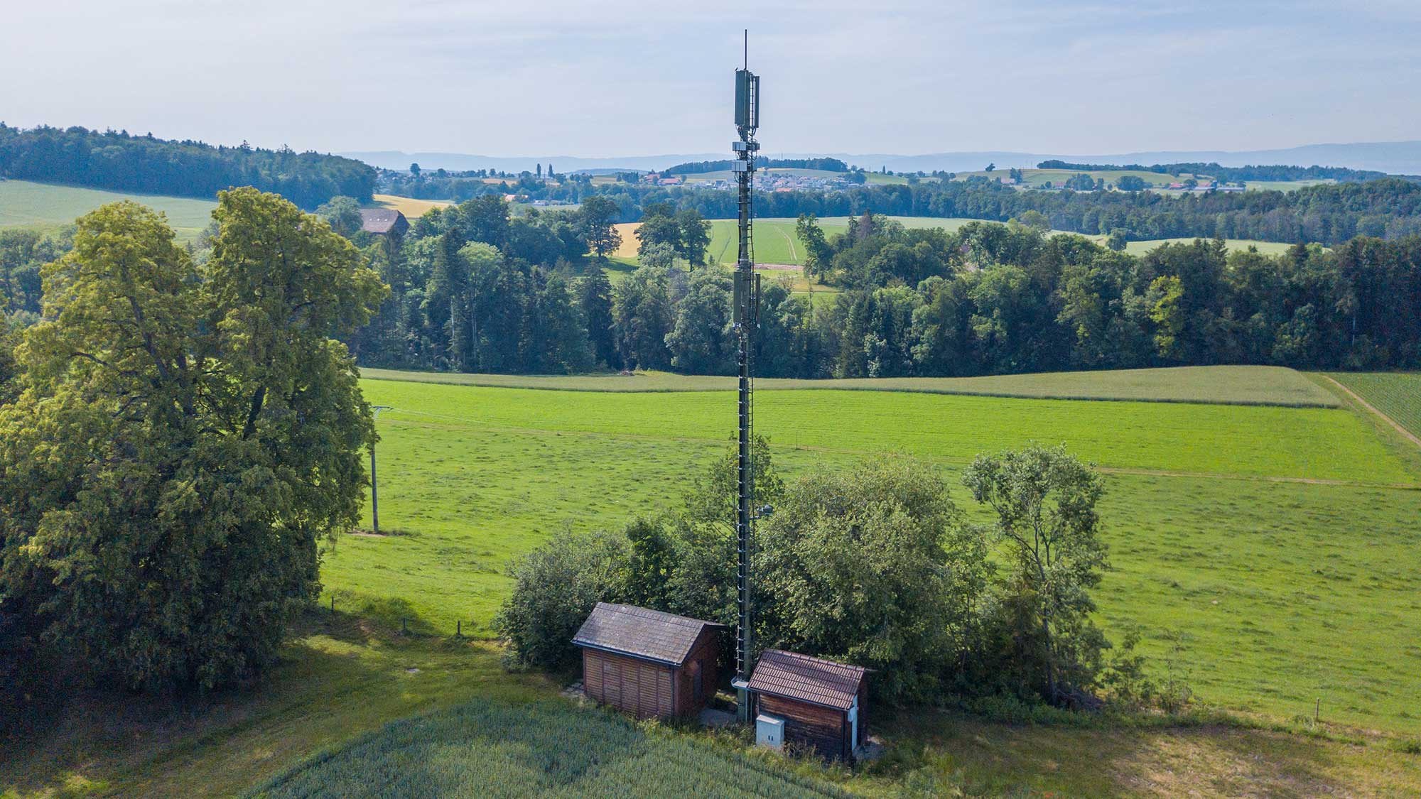 Mobilfunkmast neben zwei Häusern vor grüner Landschaft mit Bäumen. Der 5G-Betrieb gelingt ohne Umweltbelastungen.