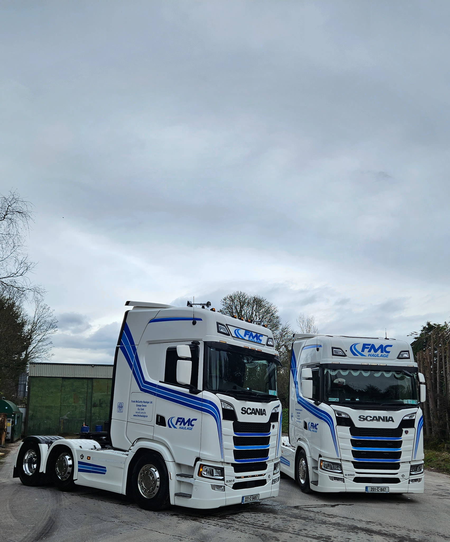 Two white Scania trucks with blue FMC Haulage livery parked side by side under a cloudy sky.
