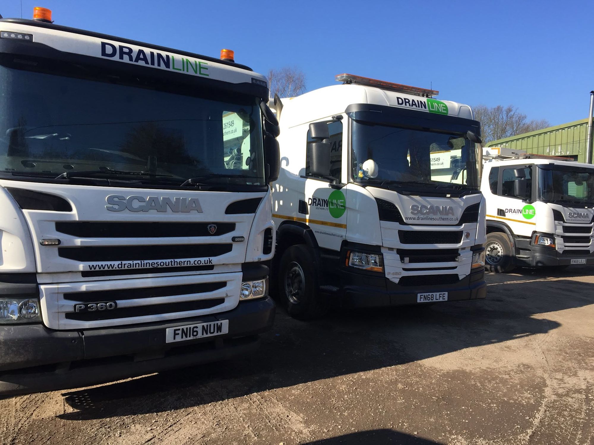Three white Scania trucks branded with Drainline Southern logos parked in a yard under a clear blue sky.