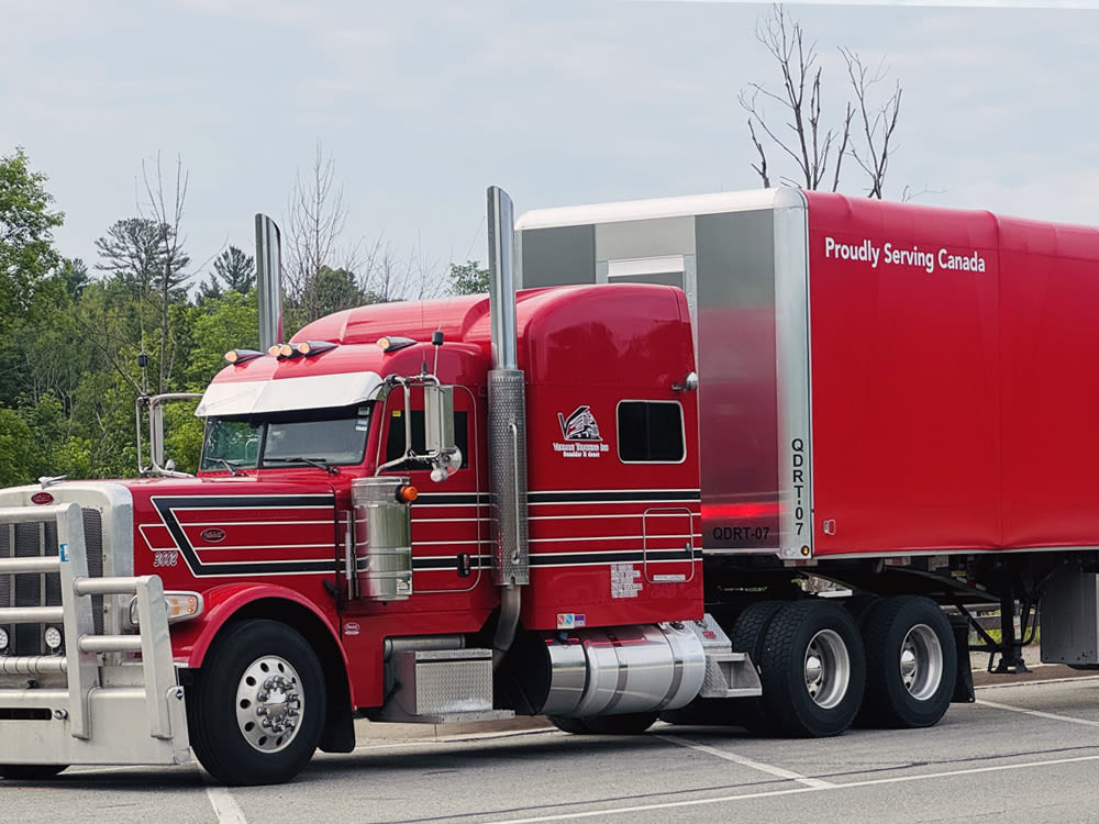 Camion semi-remorque rouge avec l’inscription « Fièrement au service du Canada », garé dans un stationnement avec des arbres.